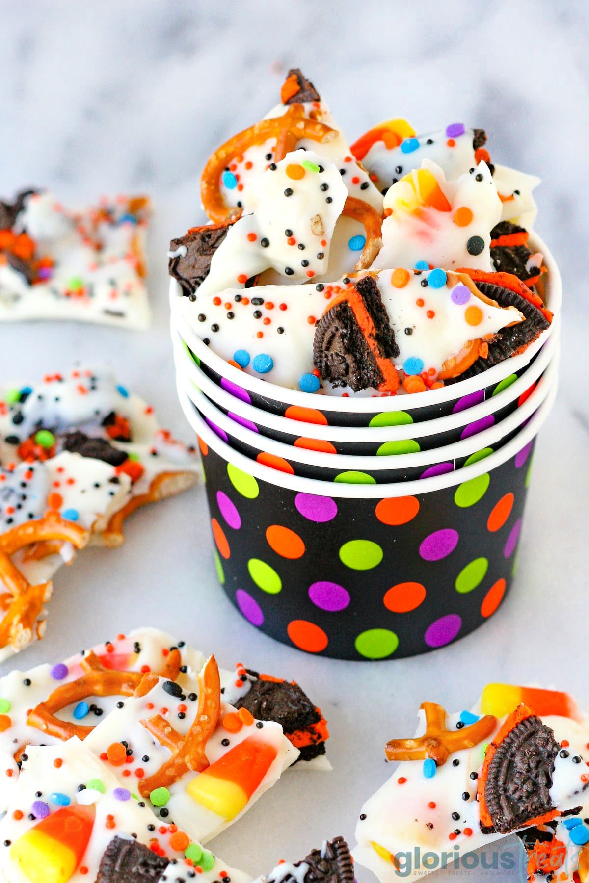 Halloween Bark broken into bite sized pieces and placed in colorful halloween cup filled to the top. Made with pretzels, sprinkles and halloween cookies and candy. More bark can be seen scatter about the base of the cup.