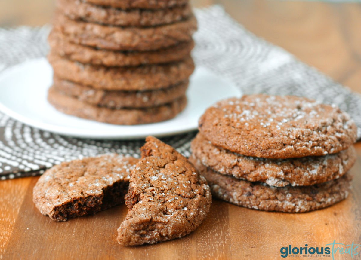 chocolate sugar cookies stacked on white plate with more cookies in the front. one cookie has been torn in half.