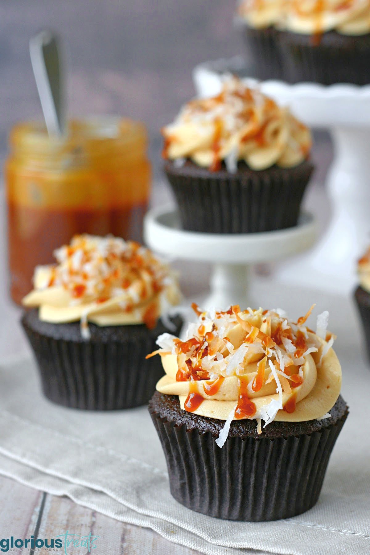 samo cupcakes artfully displayed on cupcake stands, cake stands and sitting on a beige napkin. a jar or caramel sauce with a spoon in it can be seen in the background.