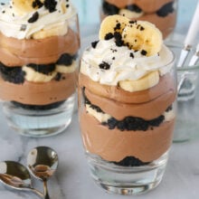 Three banana nutella trifles in glasses topped with whipped cream, OREO crumbles and fresh banana slices.