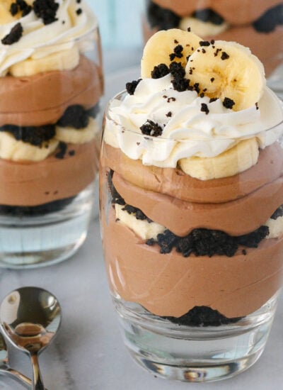 Three banana nutella trifles in glasses topped with whipped cream, OREO crumbles and fresh banana slices.