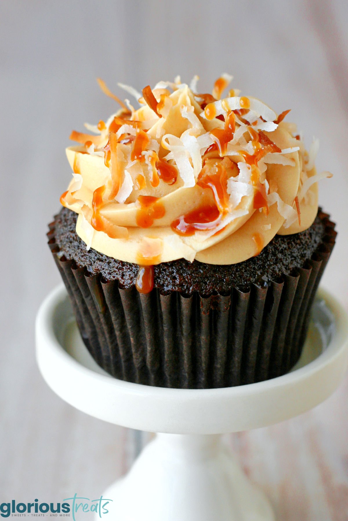 single cupcake on white cupcake stand frosted with salted caramel frosting and garnished with salted caramel sauce and toasted coconut.