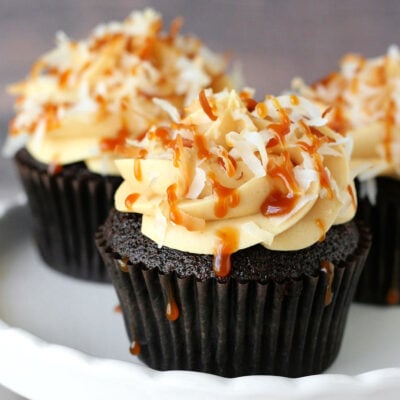 three chocolate cupcakes on a white cake stand. the cupcakes are topped with salted caramel frosting and toasted coconut creating these delicious samoa cupcakes.