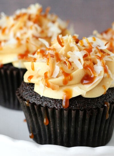 three chocolate cupcakes on a white cake stand. the cupcakes are topped with salted caramel frosting and toasted coconut creating these delicious samoa cupcakes.