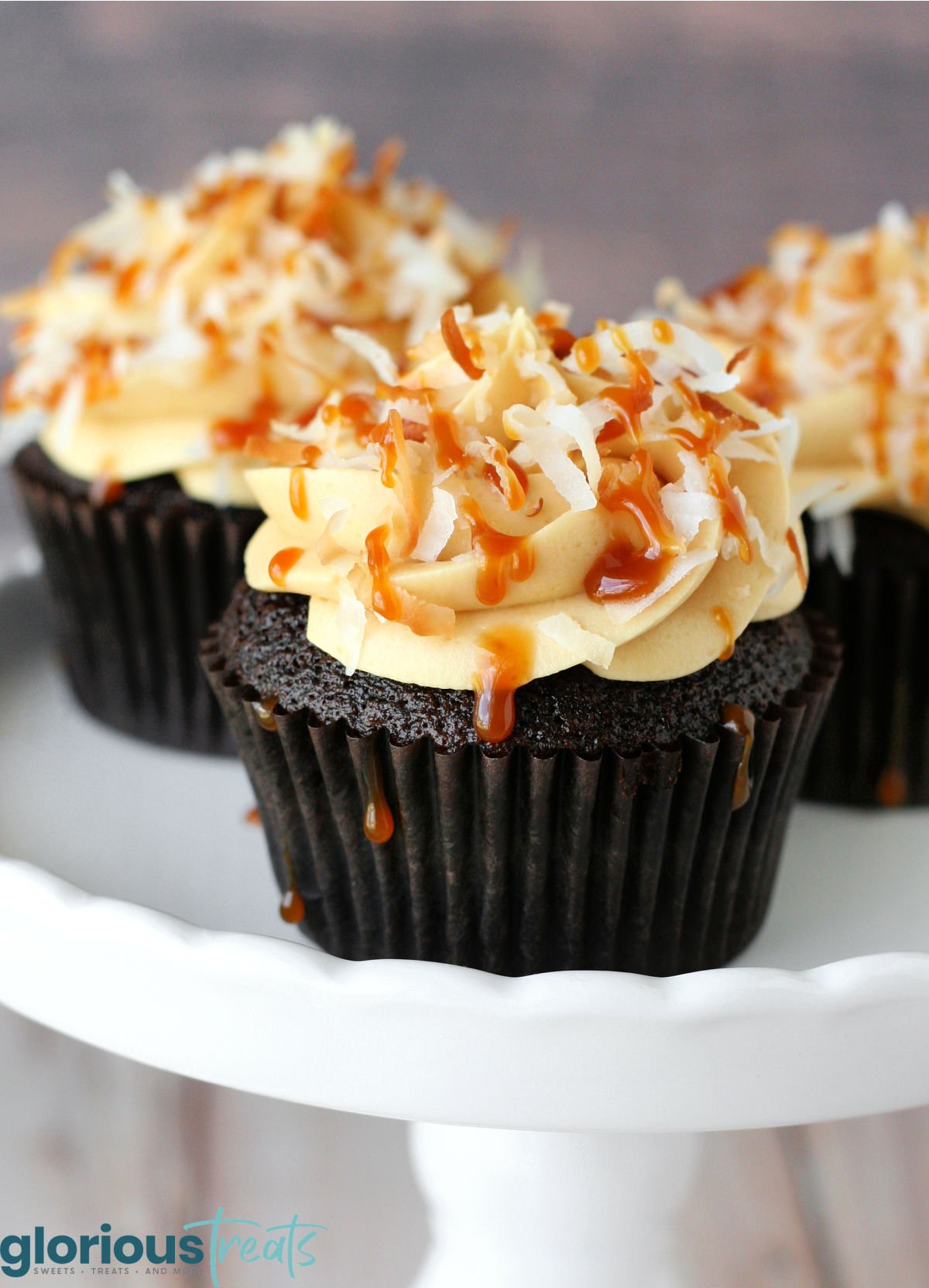 three chocolate cupcakes on a white cake stand. the cupcakes are topped with salted caramel frosting and toasted coconut creating these delicious samoa cupcakes.