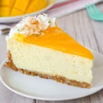 Slice of mango lime cheesecake on a round white plate. Another slices and fresh mango and limes can be seen in the background.
