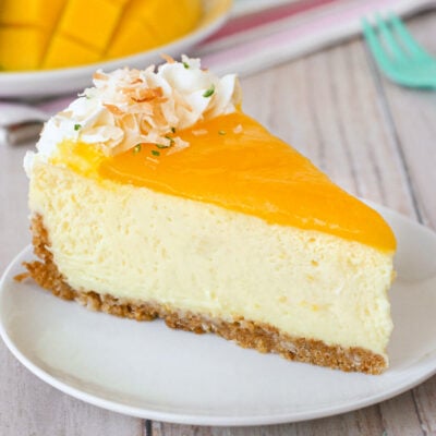 Slice of mango lime cheesecake on a round white plate. Another slices and fresh mango and limes can be seen in the background.