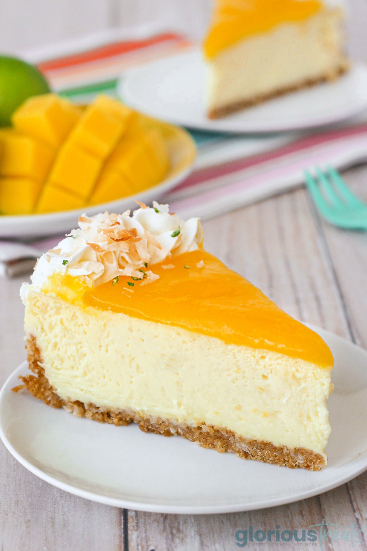 Slice of mango lime cheesecake on a round white plate. Another slices and fresh mango and limes can be seen in the background.