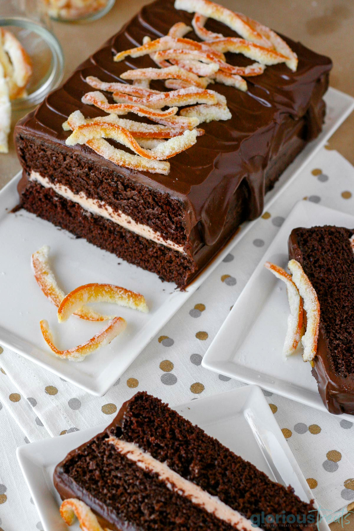 Overhead shot of chocolate orange cake recipe on white rectangular serving tray. Cake is topped with candied orange peel. Two slices have been cut and plated.