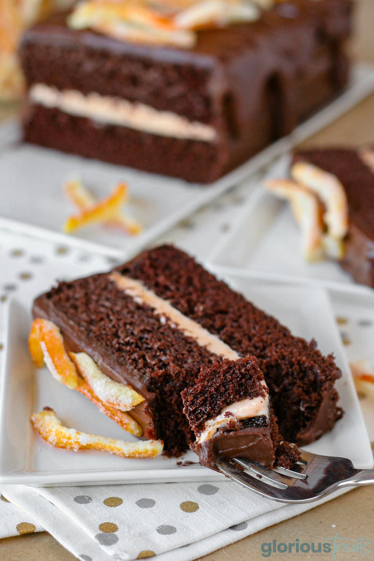 Slice of two layered chocolate orange cake on white square plate. The rest of the cake can be seen in the background.