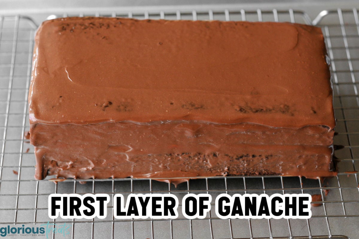 Cooled chocolate cake covered with first layer of ganache sitting on wire rack over a baking sheet.