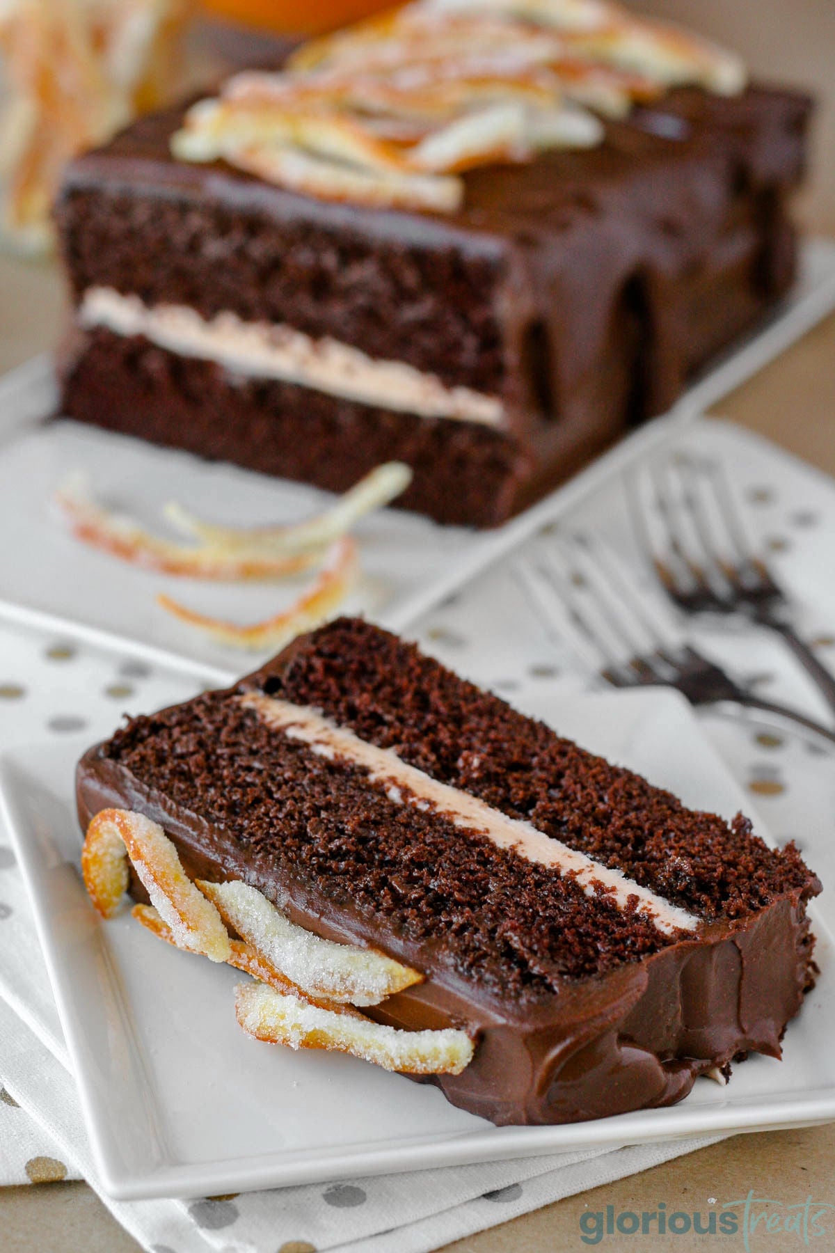 Slice of two layered chocolate orange cake on white square plate. The rest of the cake can be seen in the background.