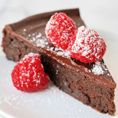 slice of flourless chocolate cake on a white round plate topped with raspberries and a dusting of powdered sugar.