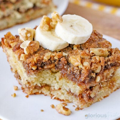 piece of banana bread coffee cake on a white round plate topped with two slices of banana. one bite has been removed.