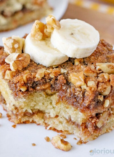 piece of banana bread coffee cake on a white round plate topped with two slices of banana. one bite has been removed.