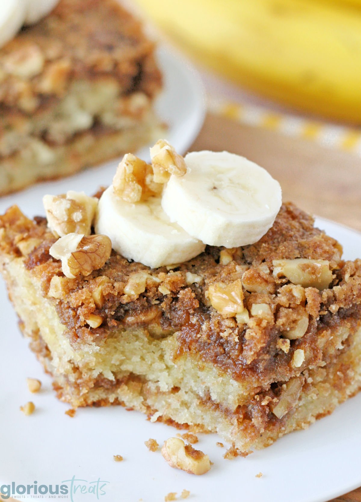 piece of banana bread coffee cake on a white round plate topped with two slices of banana. one bite has been removed.