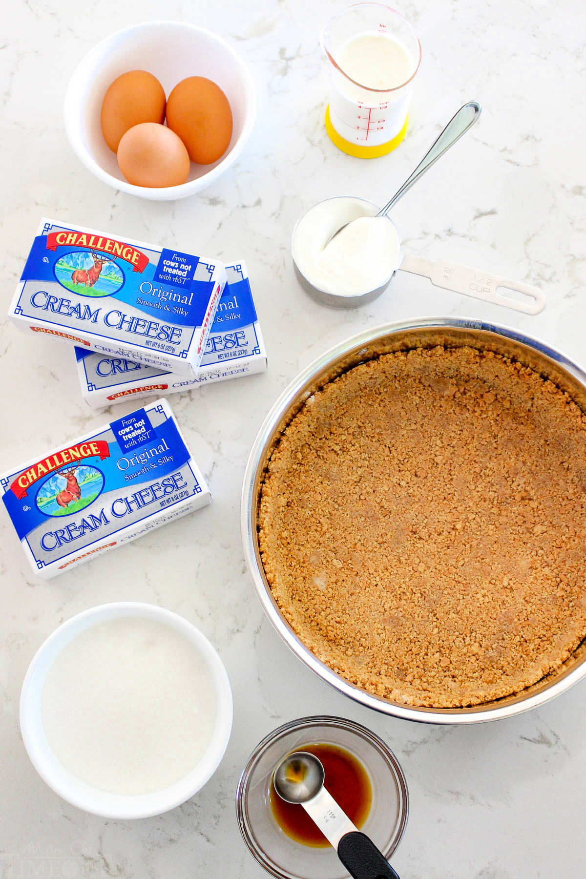 cheesecake ingredients sitting next to graham cracker crust on marble surface.