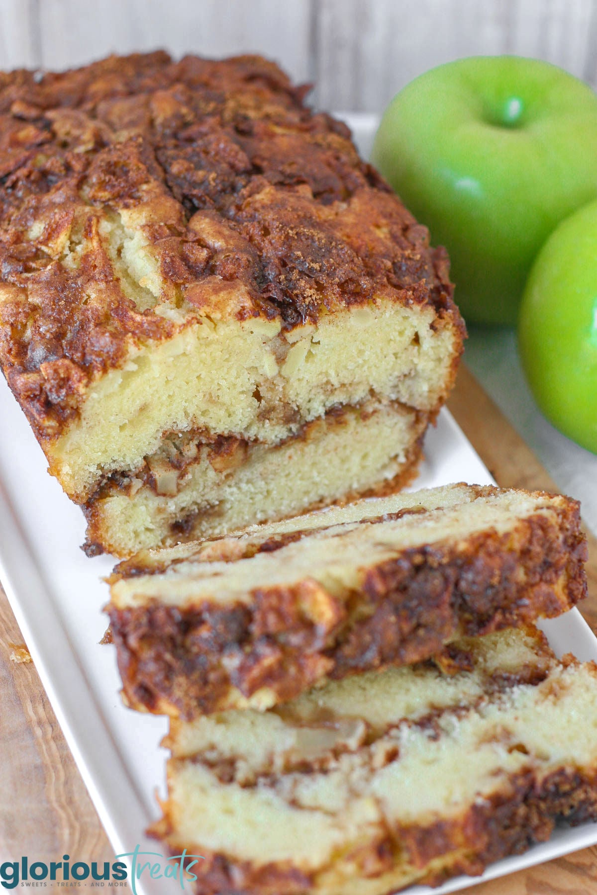 Overhead shot of loaf of apple bread on white serving tray with two slices cut. Green apples can be seen in the background.
