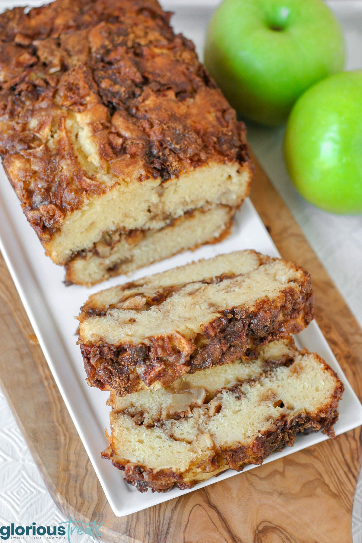 verhead shot of loaf of apple bread on white serving tray with two slices cut. Green apples can be seen in the background.
