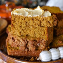 A few slices of pumpkin bread stacked on a brown hand thrown pottery plate. Top piece of pumpkin bread is slathered with butter. A decorative pumpkin themed butter knife is resting next to the bread.