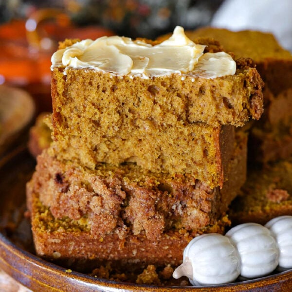 A few slices of pumpkin bread stacked on a brown hand thrown pottery plate. Top piece of pumpkin bread is slathered with butter. A decorative pumpkin themed butter knife is resting next to the bread.