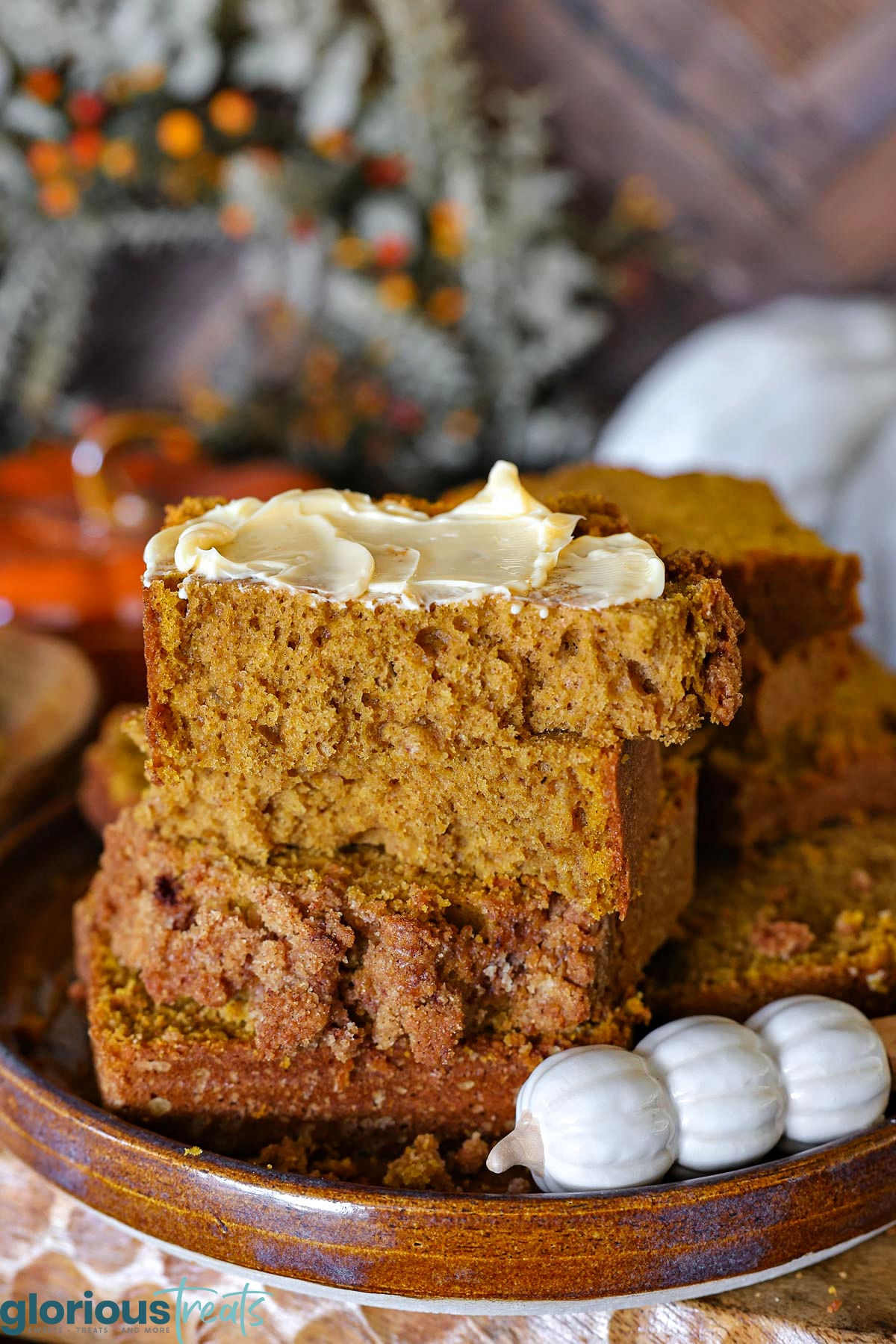 A few slices of pumpkin bread stacked on a brown hand thrown pottery plate. Top piece of pumpkin bread is slathered with butter. A decorative pumpkin themed butter knife is resting next to the bread.