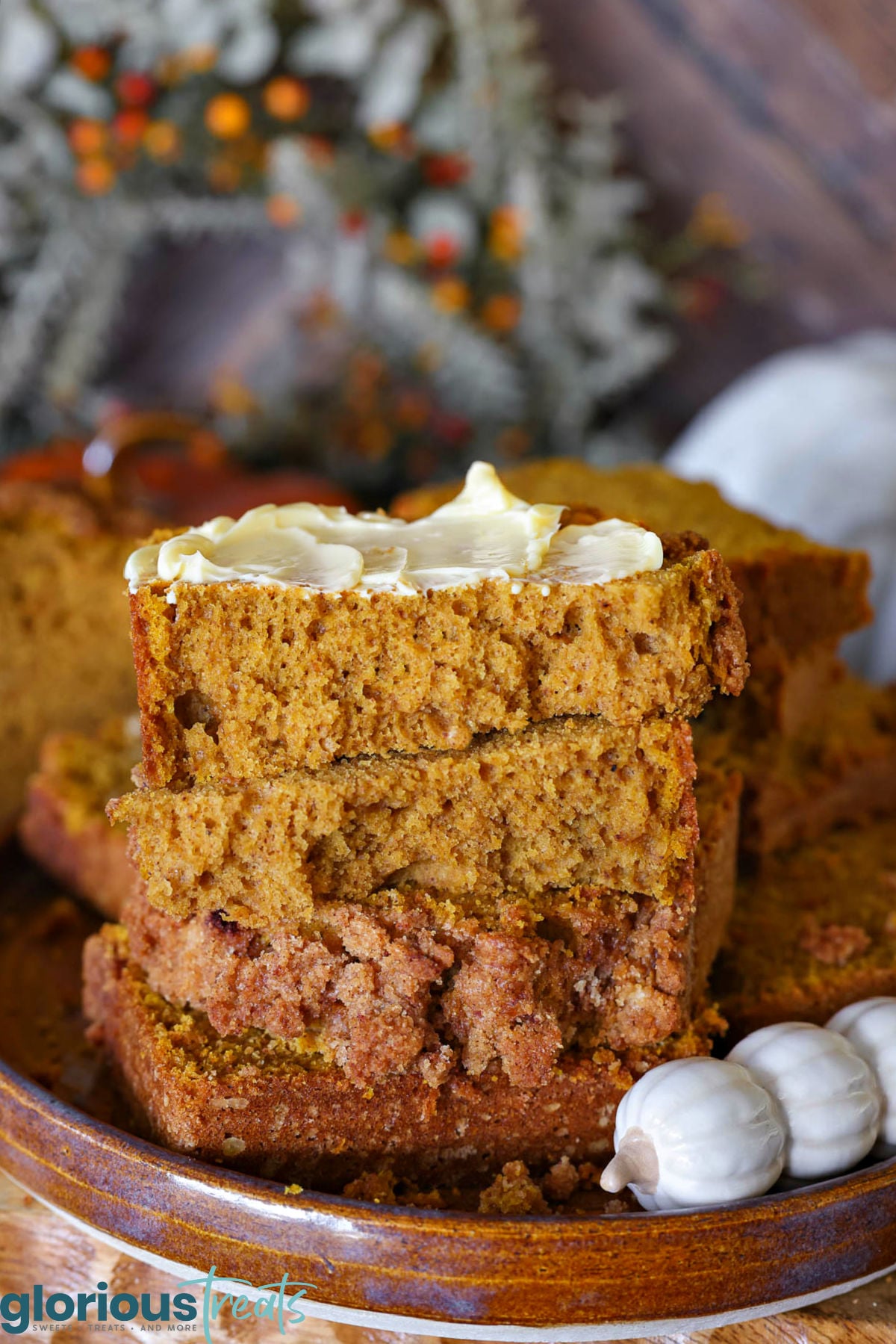 A few slices of pumpkin bread stacked on a brown hand thrown pottery plate. Top piece of pumpkin bread is slathered with butter. A decorative pumpkin themed butter knife is resting next to the bread.