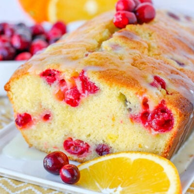 cranberry orange bread sitting on white tray with slices of oranges around the bread and fresh cranberries on the top.