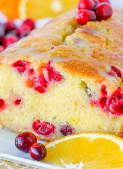 cranberry orange bread sitting on white tray with slices of oranges around the bread and fresh cranberries on the top.