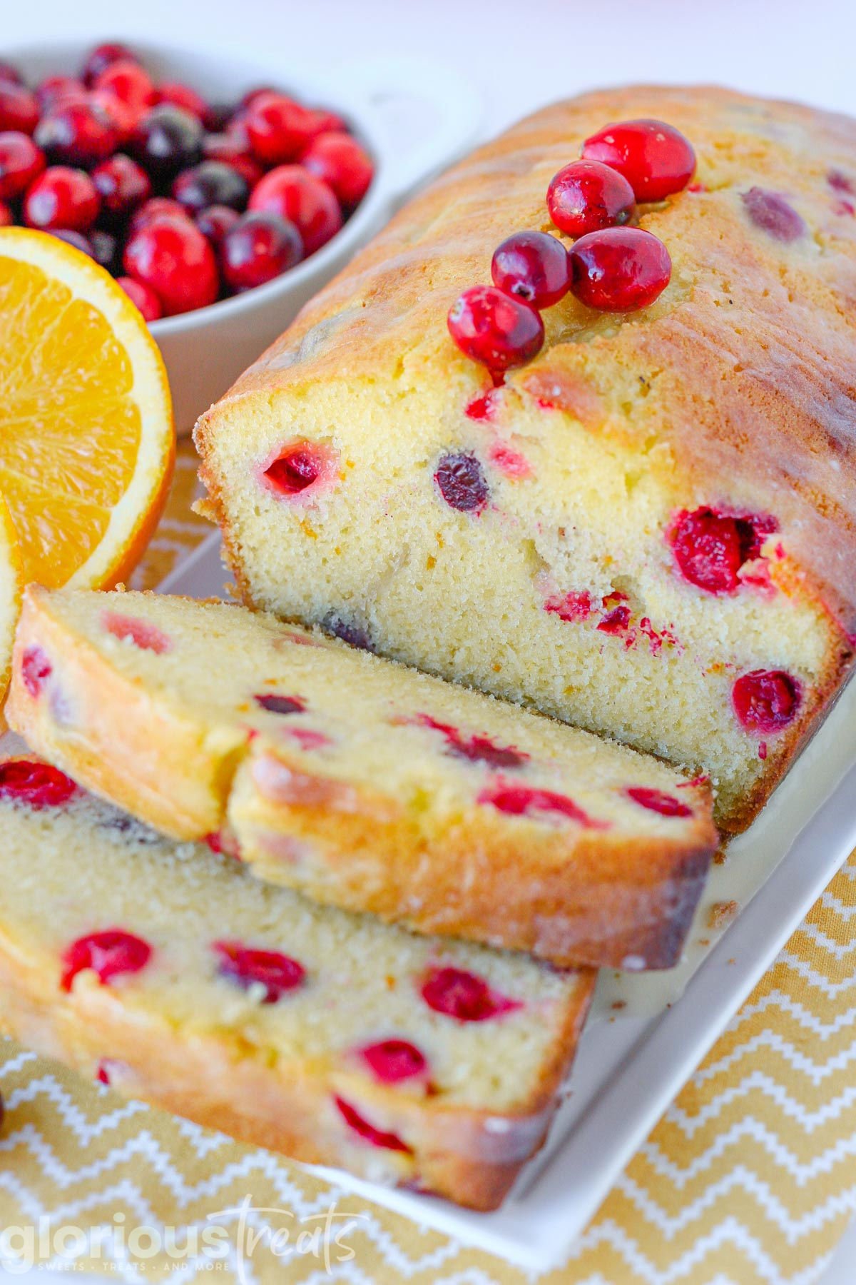 loaf of orange cranberry bread on white tray with two slices cut off. Orange wedges and fresh cranberries are to the side.