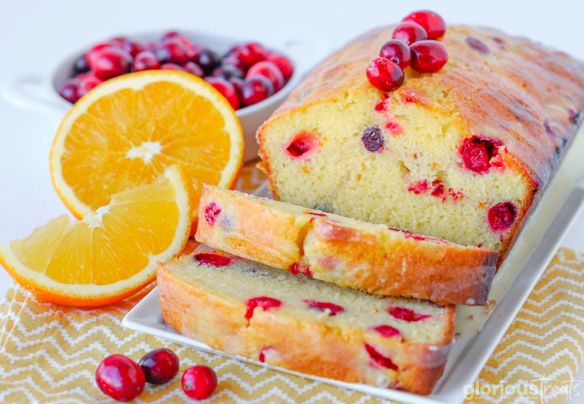 loaf of orange cranberry bread on white tray with two slices cut off. Orange wedges and fresh cranberries are to the side.
