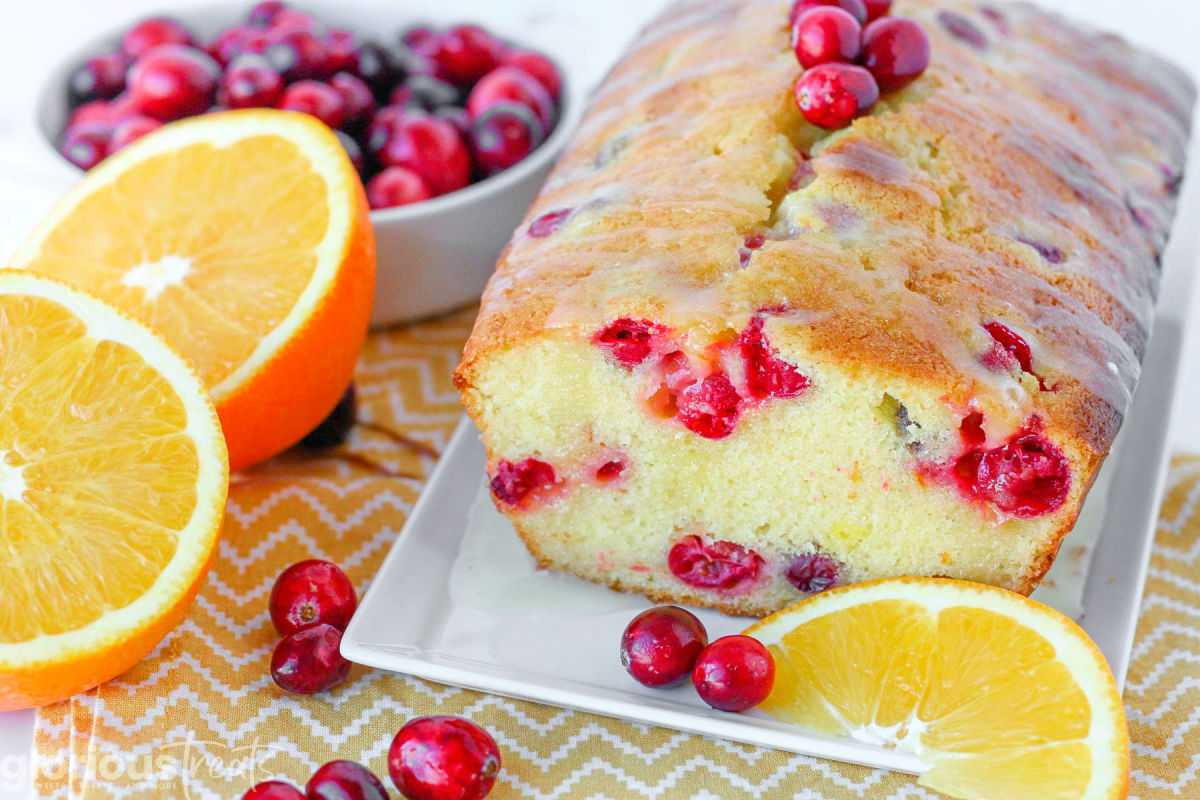 wide view of cranberry orange bread on a white glass tray with one slice removed. a bowl of fresh cranberries and orange halves can be seen next to the bread.
