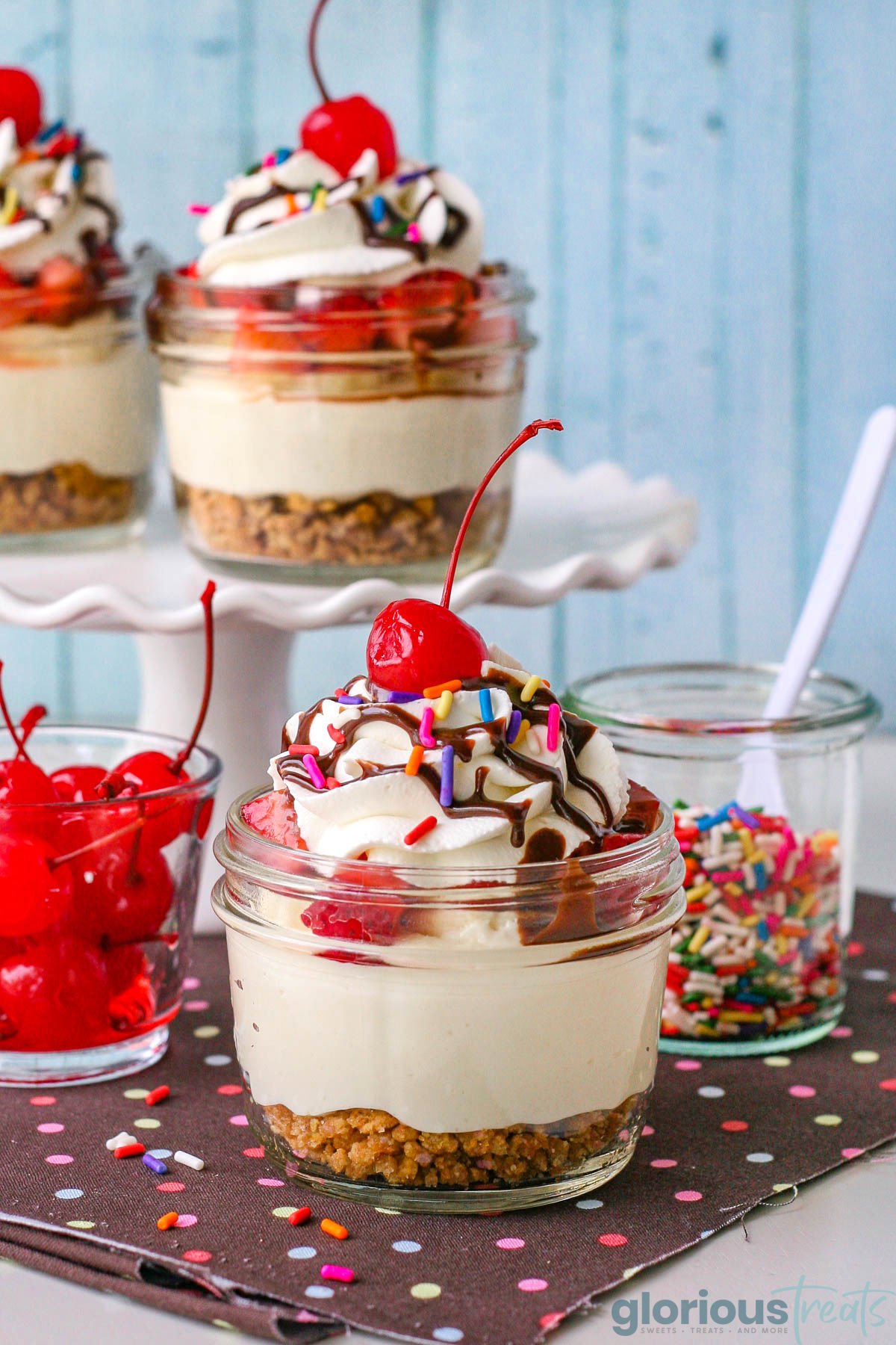 Pulled back look at banana split cheesecake in jar with two more sitting on cake stand in background.