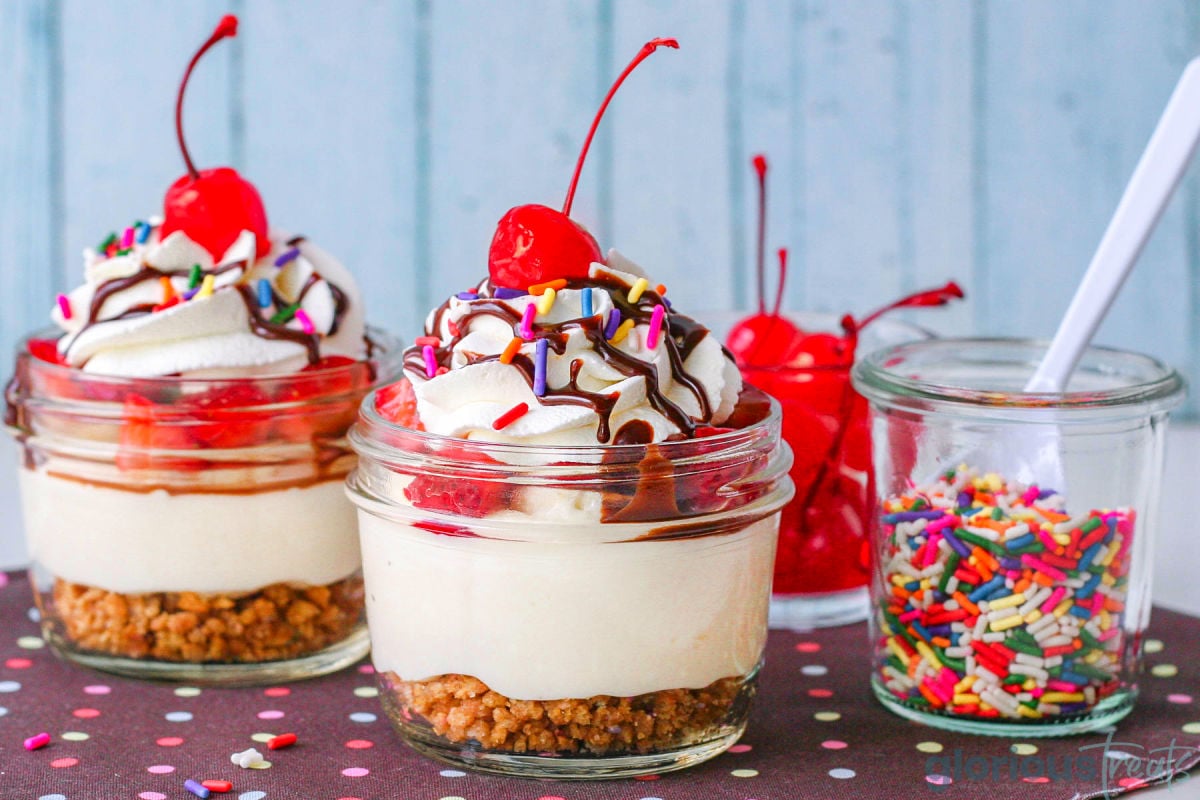 Wide shot of two no bake banana split cheesecake in jars. Topped with maraschino cherries and whipped cream.