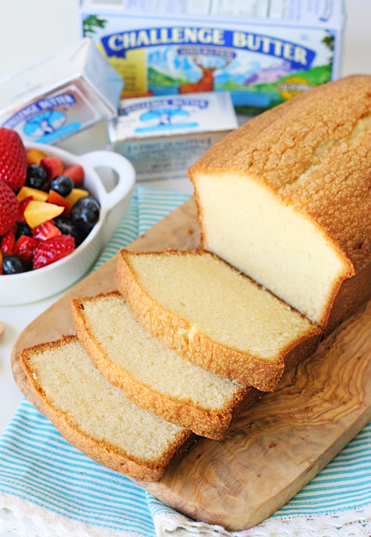 loaf pound cake on wood board with 3 slices cut sitting next to a bowl of berries.