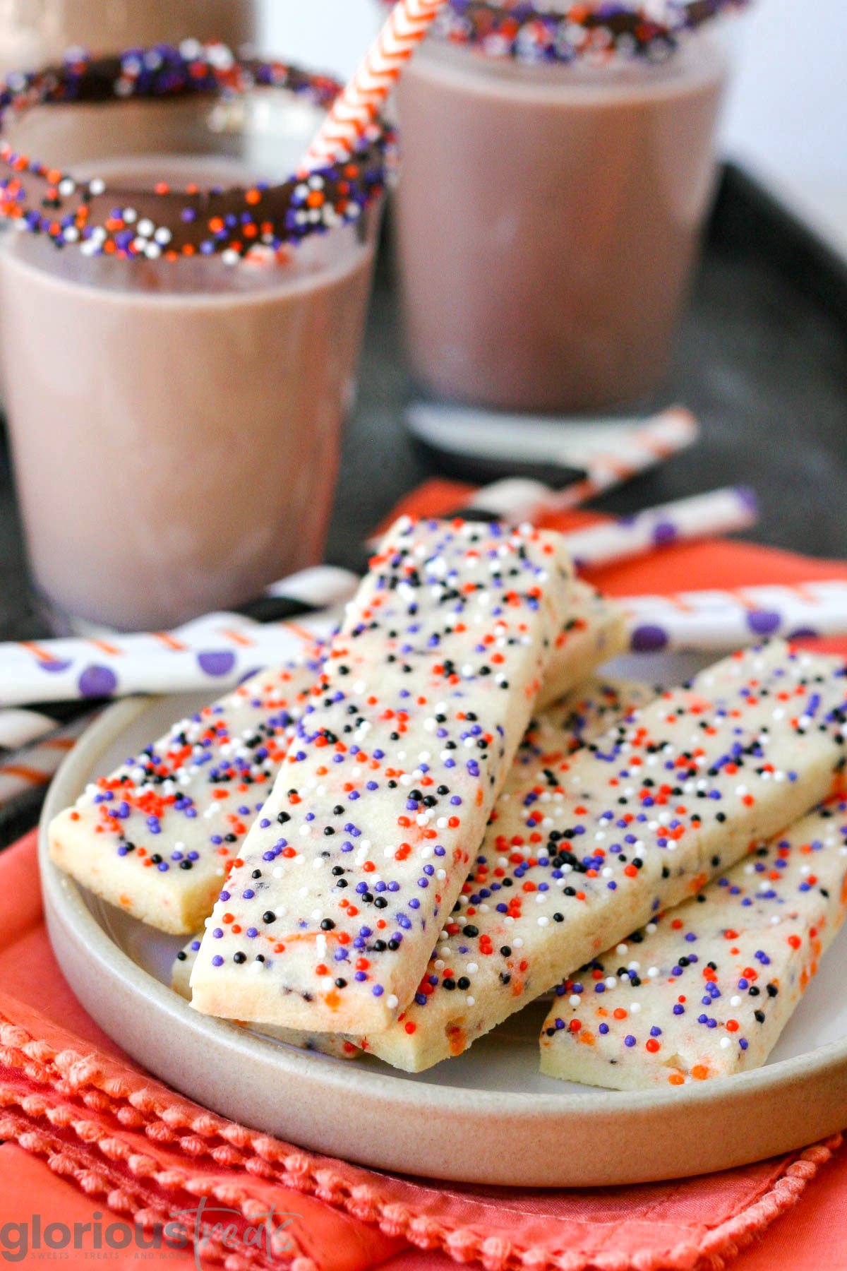 Halloween sprinkle cookies on white round plate with glasses of chocolate milk in the background. Striped paper straws are behind the cookies and the plate sits on an orange cloth napkin.