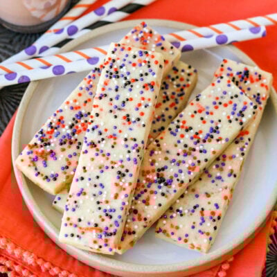 Halloween sprinkle cookies on white round plate with glasses of chocolate milk in the background. Striped paper straws are behind the cookies and the plate sits on an orange cloth napkin.