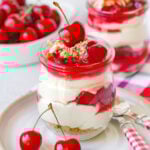A jar of no bake cherry cheesecake on a white plate with another jar and a bowl full of cherries out of focus in the background.