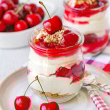 A jar of no bake cherry cheesecake on a white plate with another jar and a bowl full of cherries out of focus in the background.