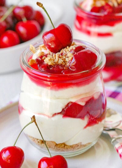 A jar of no bake cherry cheesecake on a white plate with another jar and a bowl full of cherries out of focus in the background.