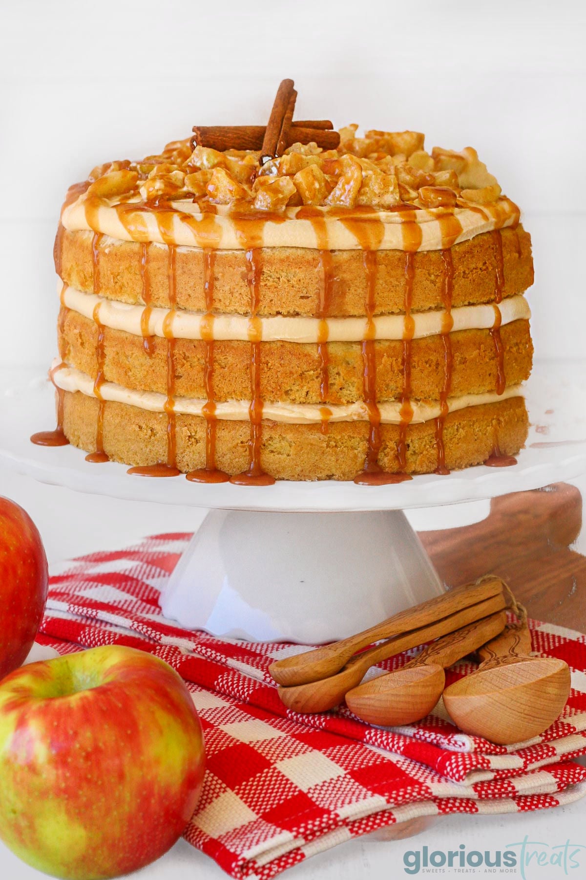 Three layer apple cake with caramel frosting and spice apple topping on white cake stand on top of red checked towel. Cake is topped with two cinnamon sticks.