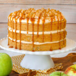 Layered caramel apple cake with caramel frosting and spiced apples on white scalloped cake stand.