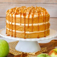 Layered caramel apple cake with caramel frosting and spiced apples on white scalloped cake stand.