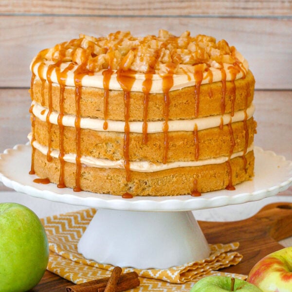 Layered caramel apple cake with caramel frosting and spiced apples on white scalloped cake stand.