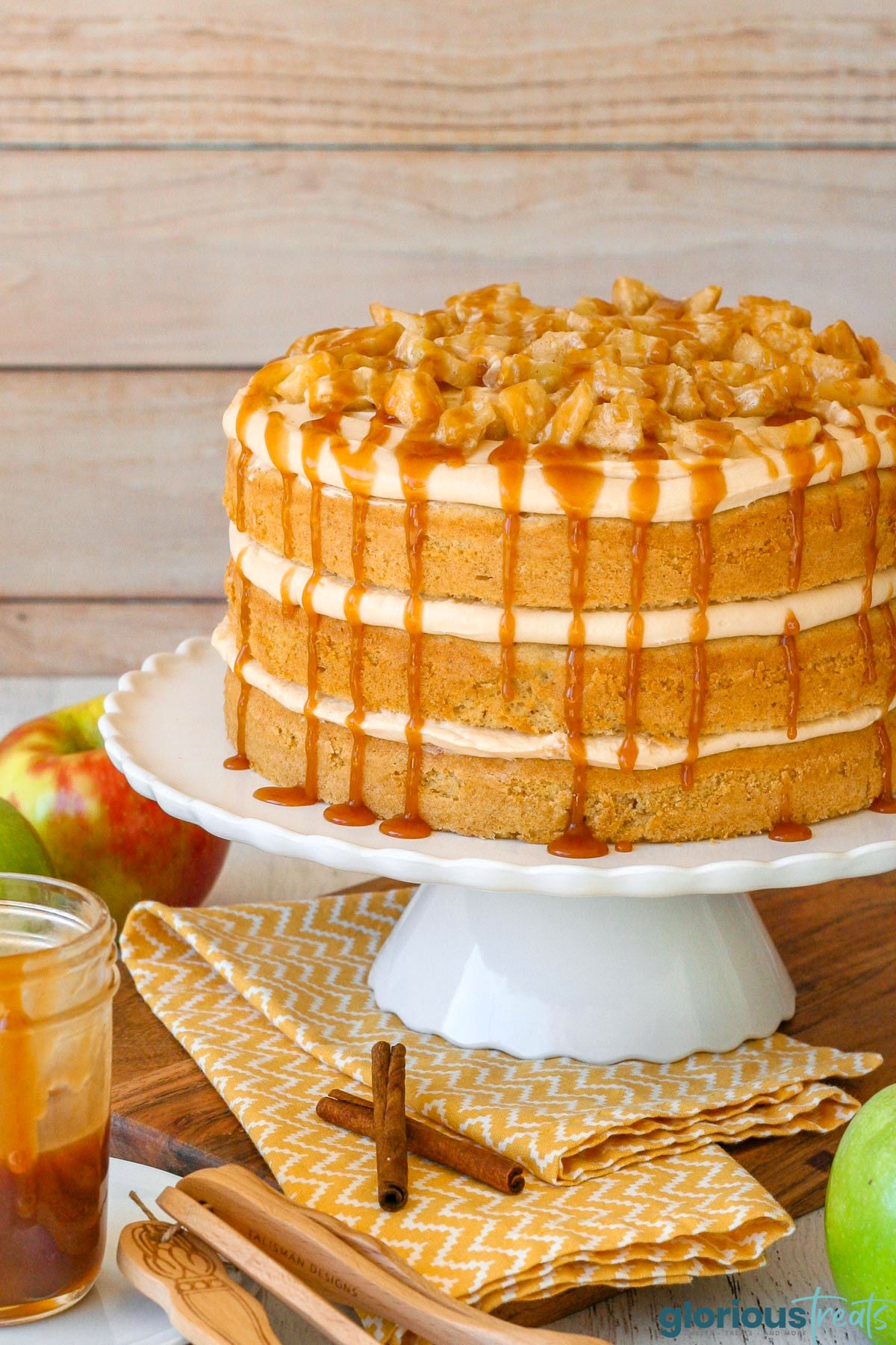 Layered caramel apple cake with caramel frosting and spiced apples on white scalloped cake stand.