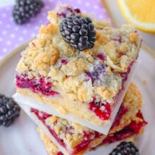 Blackberry bars stacked three high on a ceramic plate with scattered blackberries about as well as a lemon cut in half.
