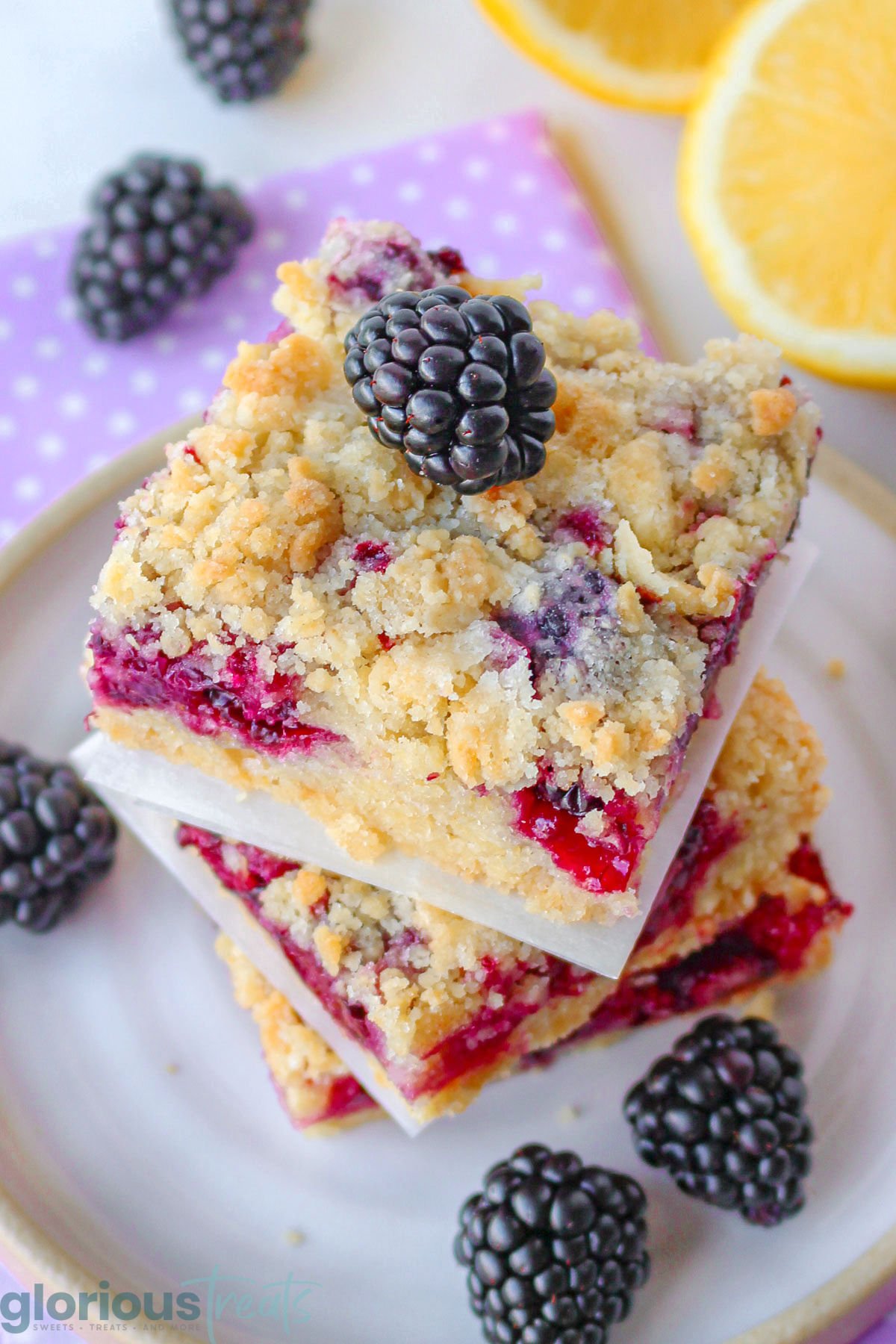 Blackberry bars stacked three high on a ceramic plate with scattered blackberries about as well as a lemon cut in half.