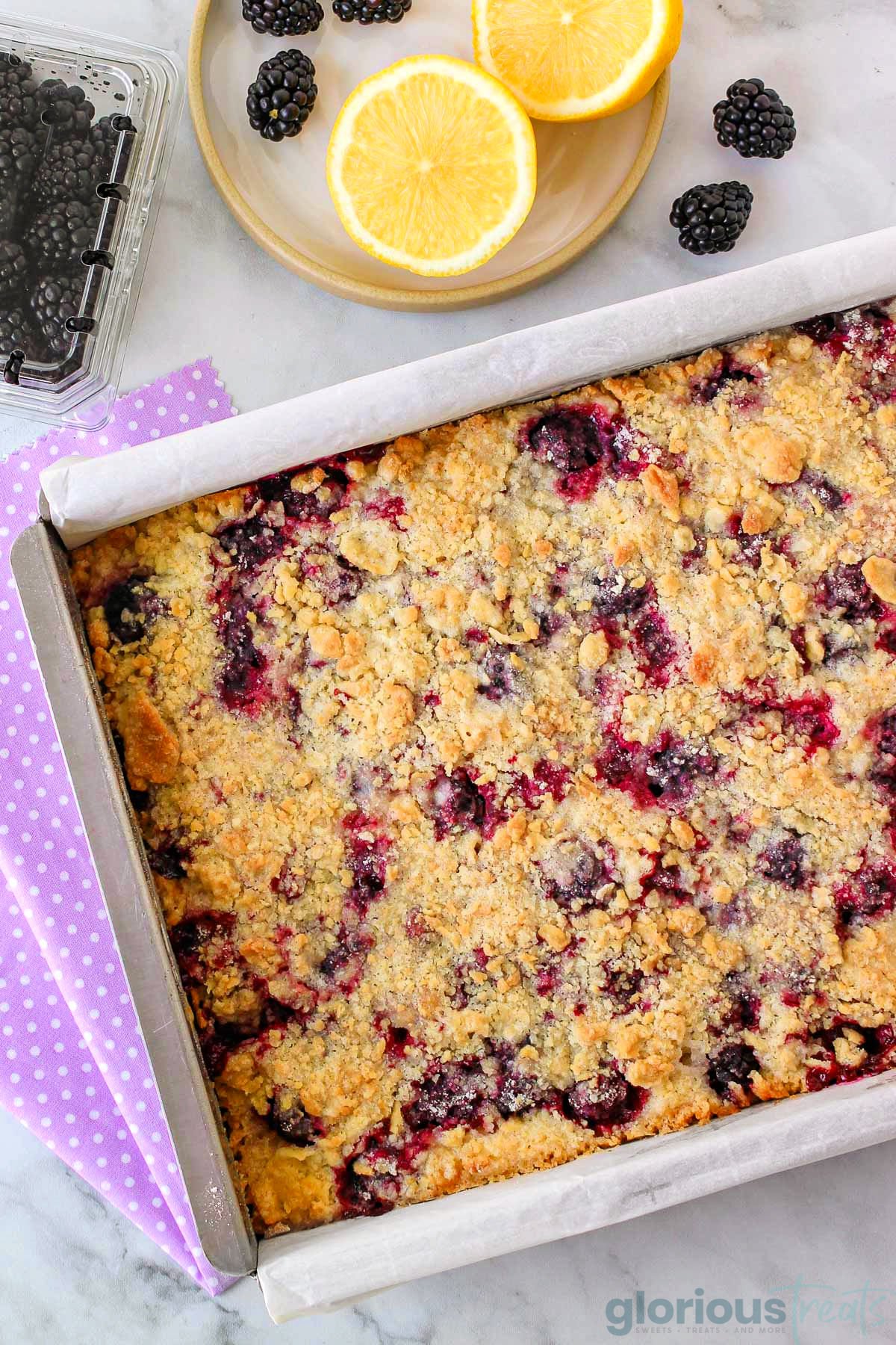 Overhead shot of pan of blackberry crumb bars at an angle next to a lemon and fresh blackberries.