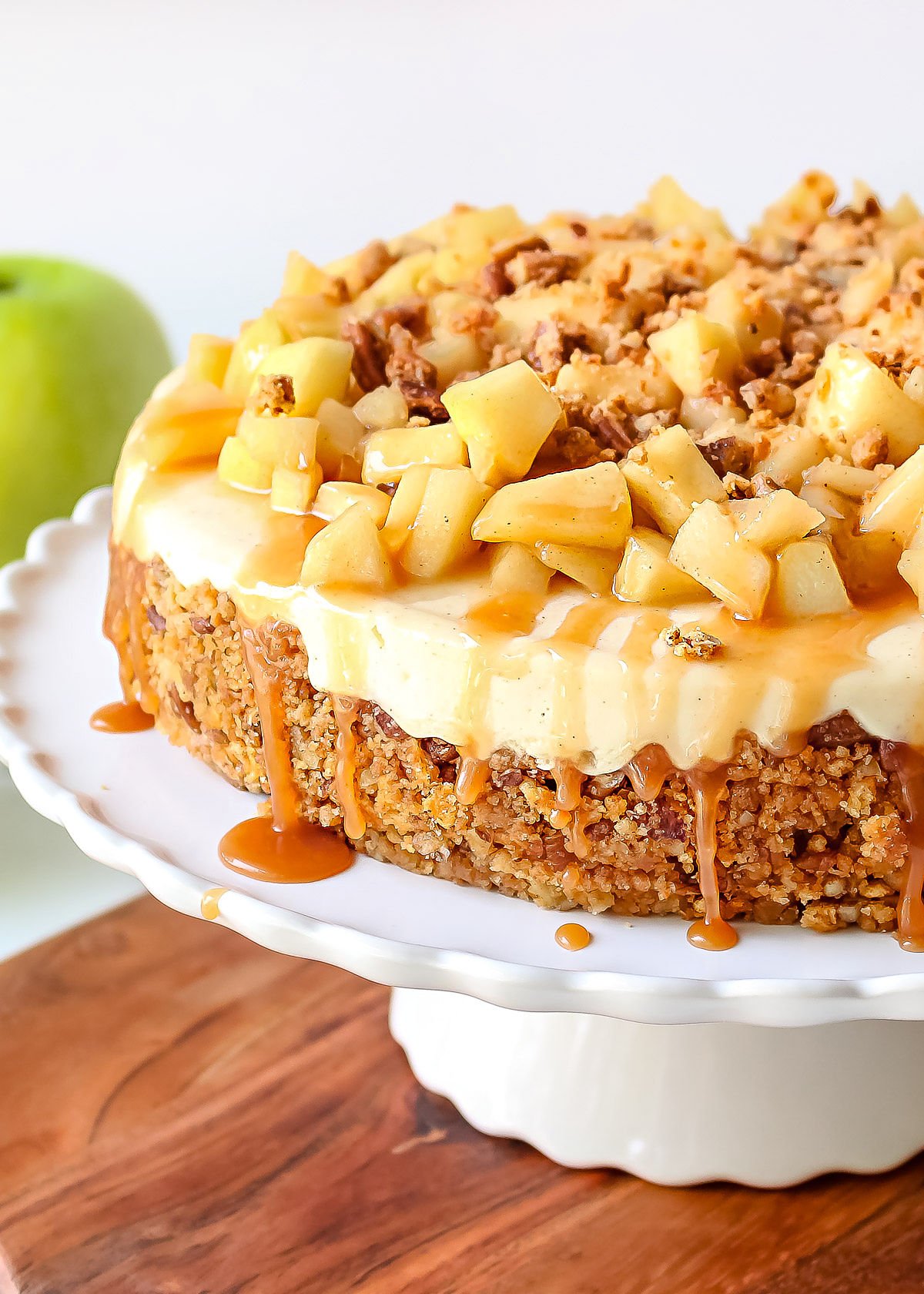 front view caramel apple cheesecake sitting on white cake stand sitting on a wood cutting board with a green apple in the background