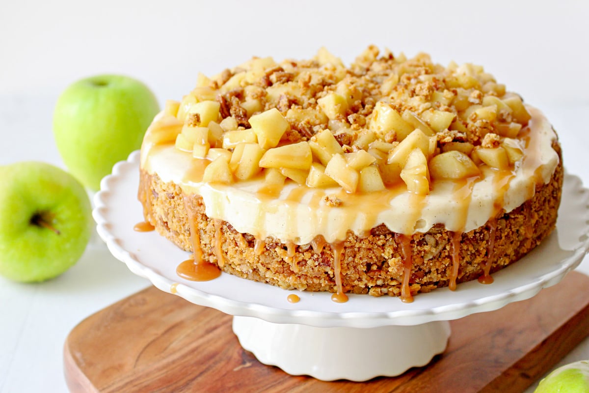 whole caramel apple cheesecake on white cake plate on wood board